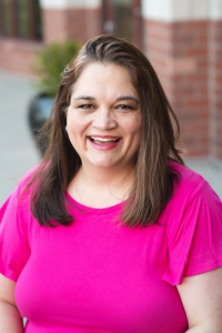 Image of a smiling white woman with dark hair in a pink shirt outside with a brick wall in the background